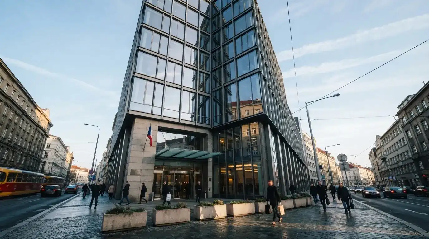Fasáda moderní administrativní budovy s skleněnými okny odráží ranní oblohu nad rušnou pražskou ulicí.